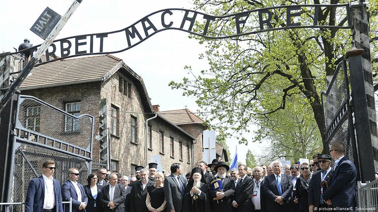 Οι επικεφαλής της «Πορείας των Ζώντων» με τον Πατριάρχη Βαρθολομαίο και των Νίκο Βούτση στην είσοδο του στρατοπέδου Άουσβιτς με το κυνικό σύνθημα «Η εργασία απελευθερώνει»