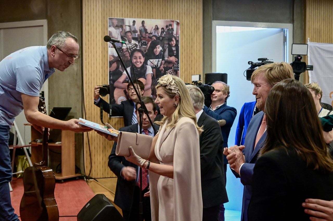 Ο Πύρρος Πουλίτσας συντονιστής της εκδήλωσης και δάσκαλος κιθάρας προσφέρει ως δώρο στην βασίλισσα το βιβλίο του Rainbow songs το οποίο είναι μέθοδος κιθάρας και καταγραφής τραγουδιών από της πατρίδες των προσφύγων